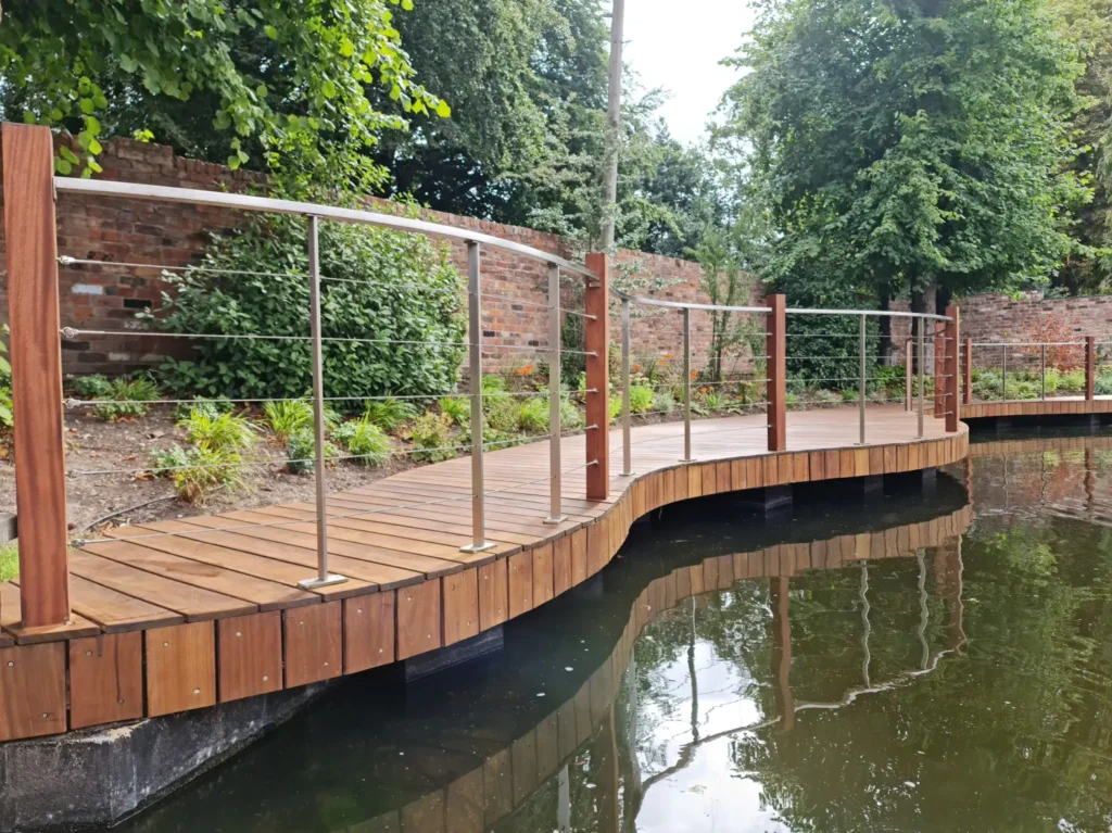 Curved timber deck bridge with stainless steel handrail beside renovated pool in Wilmslow