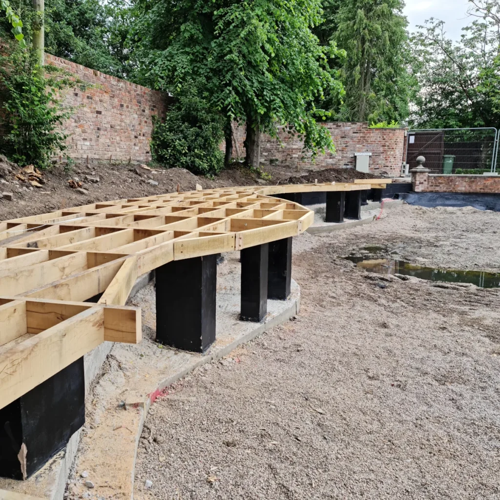 Progress of a building foundation with wooden beams and concrete supports in a garden area.