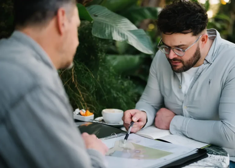 Jason and Calum discussing a project outdoors with a notebook and coffee.
